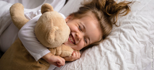 Mädchen liegt mit einem Teddybär im Bett und drückt ihn mit geschlossenen Augen ganz fest.