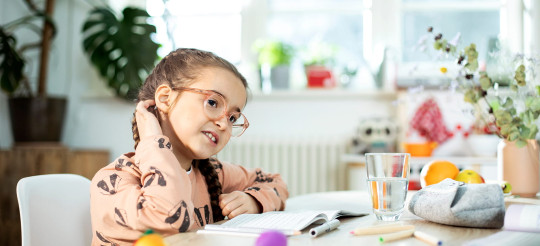 Mädchen mit Brille kratzt sich am Hinterkopf, während es an einem Tisch mit Schulheft sitzt.
