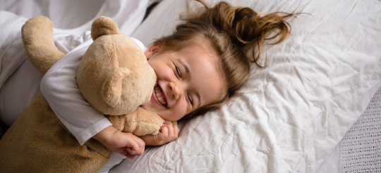 Mädchen liegt mit einem Teddybär im Bett und drückt ihn mit geschlossenen Augen ganz fest.