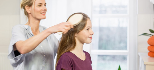 Frau kämmt mir einer Haarbürste die langen Haare eines sitzenden Mädchens. 