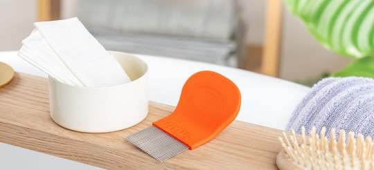Orangefarbener Läuse- und Nissenkamm neben weißen Tüchern und Haarbürste auf einem Holzbrett.