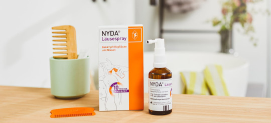 NYDA Läusespray mit Packung, Pumpflasche und orangefarbenem Kamm auf einem hellen Holztisch.