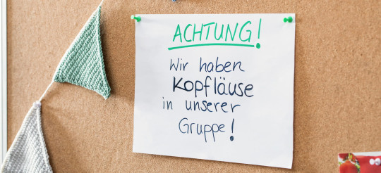 Notizzettel „Achtung! Wir haben Kopfläuse in unserer Gruppe!“ hängt mit Nadeln an einer Pinnwand.
