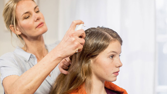 Frau sprüht NYDA Läusespray auf den Scheitel eines sitzenden Mädchens mit langen Haaren.