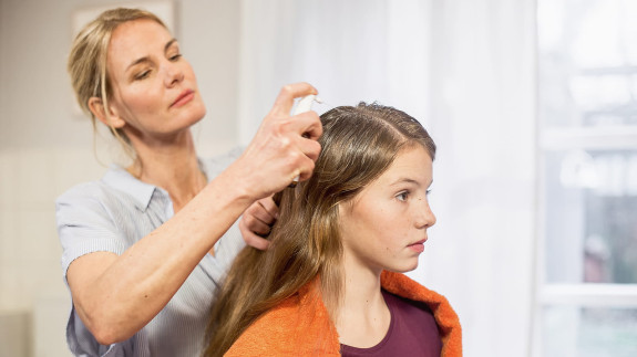 Frau sprüht NYDA Läusespray auf den Scheitel eines sitzenden Mädchens mit langen Haaren.