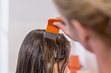 Ein Mädchen sitzt am Tisch und kämmt mit einem Lice-Comb durch ihr Haar. Ein Notizbuch und Spielzeug stehen vor ihr.