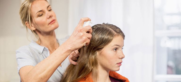 Eine Frau sprüht ein Mittel gegen Kopfläuse auf das Haar eines Mädchens, das aufmerksam zuschaut.