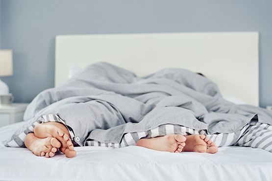 Person liegt unter einer grauen Bettdecke; nur die Hände und Füße ragen am Bettrand hervor.