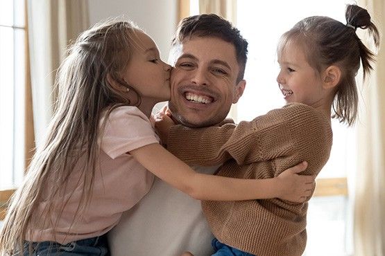 Vater sitzt mit zwei Kindern auf einem Sofa; beide Kinder umarmen und küssen ihn.