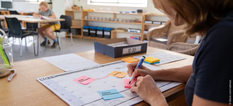 Lehrkraft sitzt im Klassenraum am Schreibtisch und schreibt Notizzettel, während Kinder im Hintergrund sitzen.