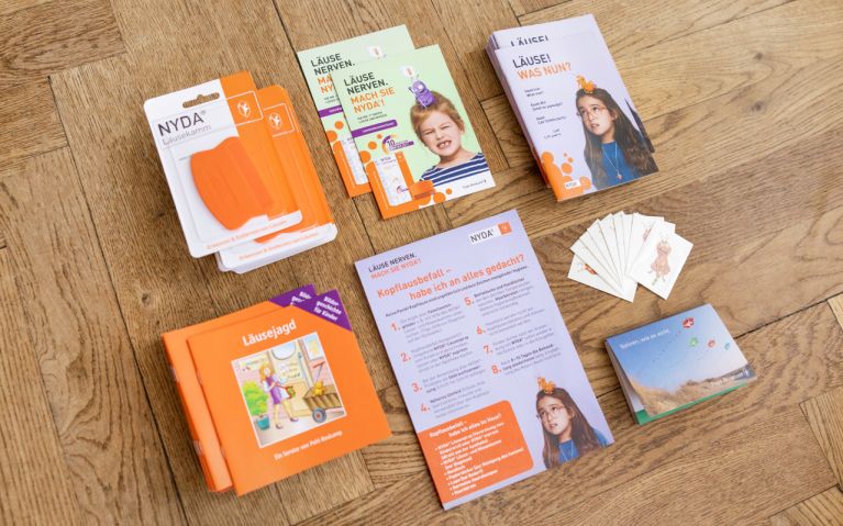 NYDA® Läusekamm in orange auf Verpackung sowie Informationsblätter und Broschüren zu Läusebefall auf Holzuntergrund.