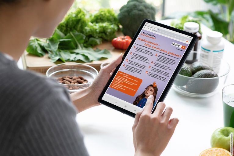 Person hält ein Tablet mit Informationen zu Läusebefall in einer Küche, frisches Gemüse im Hintergrund.