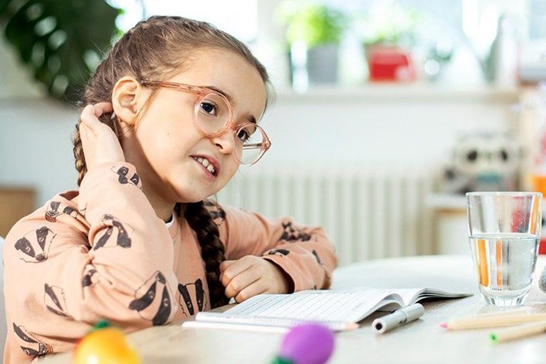 Ein Mädchen mit Brille sitzt an einem Tisch, berührt ihren Hinterkopf und lächelt. Vor ihr liegt ein Notizbuch und ein Glas.