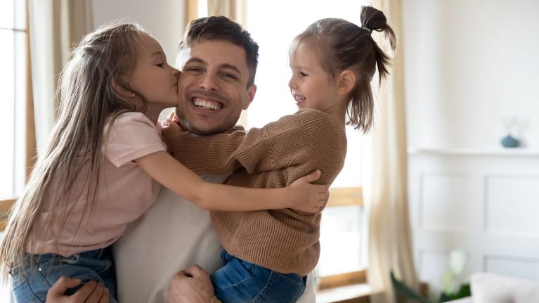 Vater sitzt mit zwei Kindern auf einem Sofa; beide Kinder umarmen und küssen ihn.