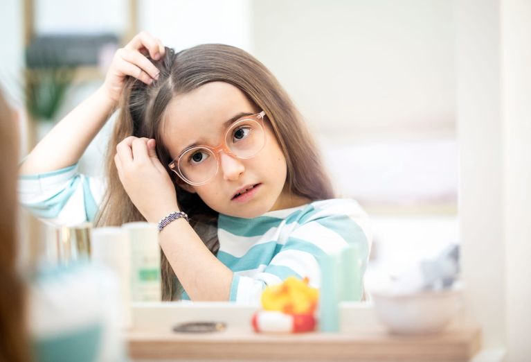 Mädchen mit Brille zieht eine Haarsträhne auseinander und kontrolliert seinen Kopf im Spiegel.