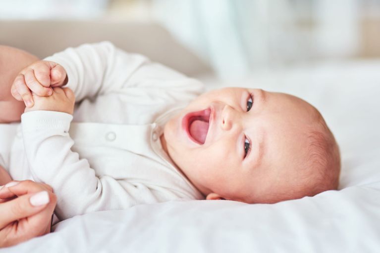 Lachendes Baby liegt auf einer weißen Decke und hält mit beiden Händen die Finger einer erwachsenen Person.