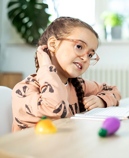 Mädchen mit Zopf und Brille sitzt am Tisch und sieht lächelnd auf ein Heft, bunte Spielzeuge liegen vor ihr.
