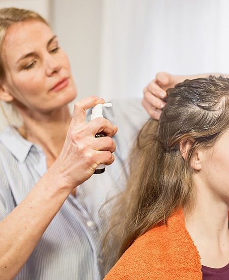 Frau sprüht Läusespray auf die Kopfhaut einer sitzenden Frau mit Handtuch um den Hals.
