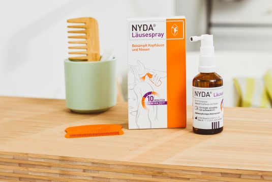 NYDA Läusespray mit Pipette und Kamm auf Holzunterlage neben grünem Behälter.