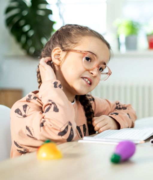 Mädchen mit Brille und Zöpfen streicht sich nachdenklich über den Kopf, sitzt an einem Tisch mit Spielzeug und Notizbuch.