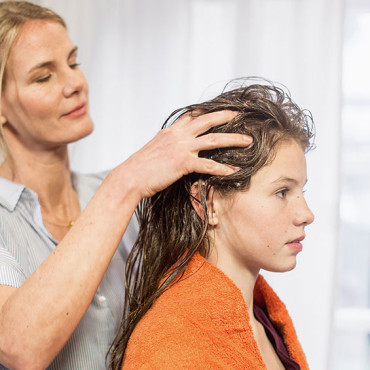 Frau massiert das Läusespray mit beiden Händen in das nasse Haar einer jungen Frau ein.
