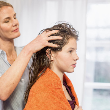 Frau massiert das Läusespray mit beiden Händen in das nasse Haar einer jungen Frau ein.