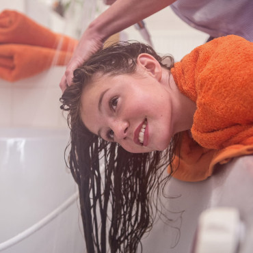 Mädchen beugt sich mit nassen Haaren über eine Badewanne und spült die Behandlung aus. 