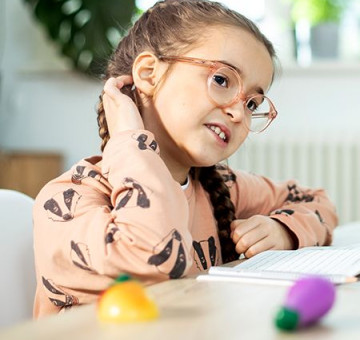 Mädchen mit Brille kratzt sich am Hinterkopf, während es an einem Tisch mit Schulheft sitzt.