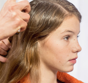 Frau sprüht NYDA Läusespray auf den Scheitel eines sitzenden Mädchens mit langen Haaren.