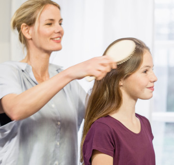 Frau kämmt mir einer Haarbürste die langen Haare eines sitzenden Mädchens. 