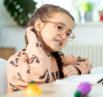 Mädchen mit Brille kratzt sich am Hinterkopf, während es an einem Tisch mit Schulheft sitzt.