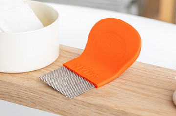 Orangefarbener Läuse- und Nissenkamm neben weißen Tüchern und Haarbürste auf einem Holzbrett.