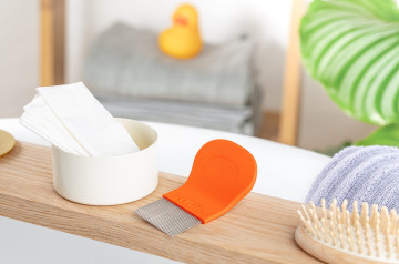 Orangefarbener Läuse- und Nissenkamm neben weißen Tüchern und Haarbürste auf einem Holzbrett.