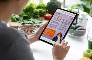 Hand hält ein Tablet mit geöffneter NYDA-Kopflaus-Checkliste über einem Küchentisch mit Lebensmitteln.