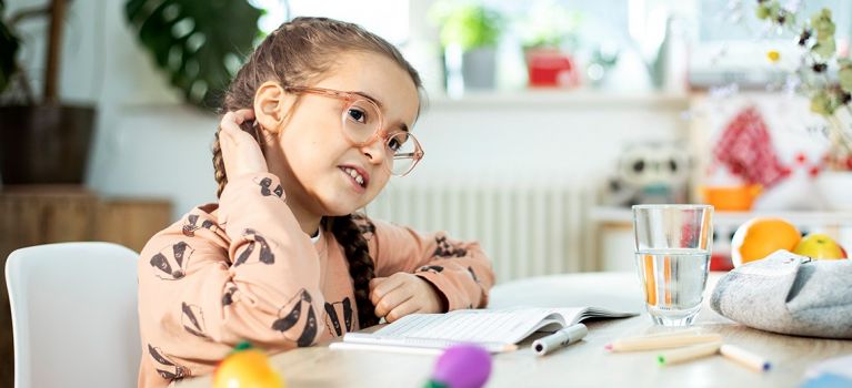 Juckreiz bei Kopfläusen – Kind kratzt sich am Kopf, mögliche Anzeichen eines Läusebefalls. Ein Mädchen mit Brille und geflochtenen Zöpfen sitzt an einem Tisch und kratzt sich am Hinterkopf. Vor ihr liegen ein Notizbuch und bunte Spielzeuge. Im Hintergrund sind Pflanzen und ein helles Fenster zu sehen.