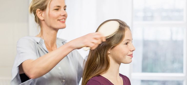 ine Frau bürstet die langen Haare eines Mädchens, das entspannt lächelt. Beide befinden sich in einem hellen Raum, und die Haare des Mädchens sind glatt und gepflegt.