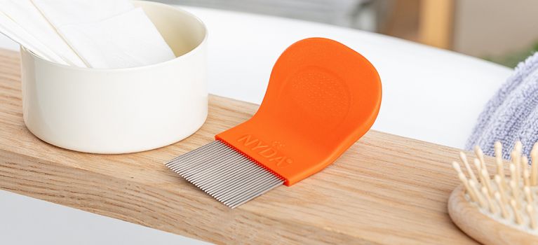 Orangefarbener NYDA® Läuse- und Nissenkamm liegt neben einem Becher auf einer Holzablage, umgeben von weiteren Pflegeutensilien.