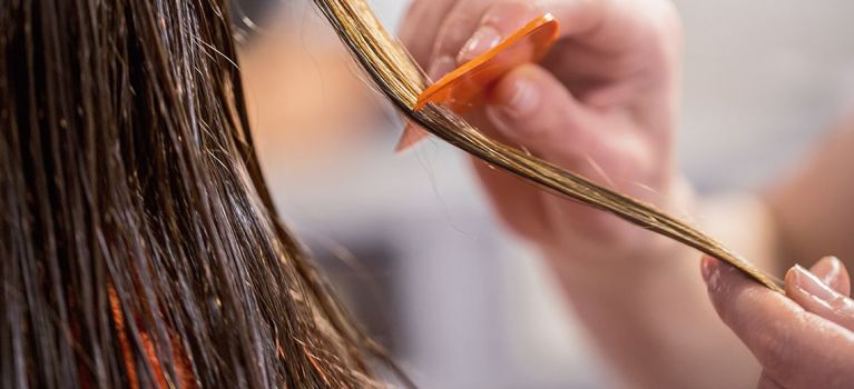 Eine Hand kämmt eine nasse Haarsträhne mit einem orangefarbenen Läusekamm aus. Der Fokus liegt auf der Strähne und dem Kamm.