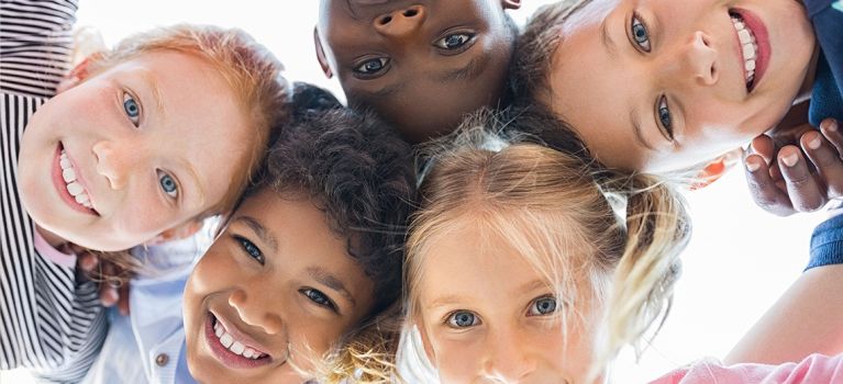 Fünf lachende Kinder verschiedener Herkunft stehen in einem Kreis und schauen fröhlich in die Kamera. Sie haben die Köpfe nah beieinander, sodass ihre Haare sich fast berühren.