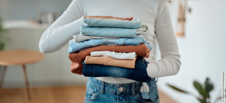 Gestapelte Kleidung in verschiedenen Farben und Stoffen, von einer Person mit weißen Ärmel aus gehalten.
