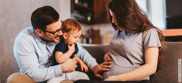 Mann sitzt mit Kleinkind auf Sofa und zeigt auf den Babybauch einer schwangeren Frau. Alle blicken lächelnd.