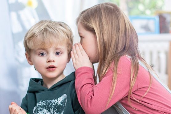 Ein blondes kleines Kind in einem dunkelgrünen Pullover schaut erstaunt, während ein Mädchen mit langen braunen Haaren ihm ins Ohr flüstert. Beide Kinder sitzen in einem hellen Raum.