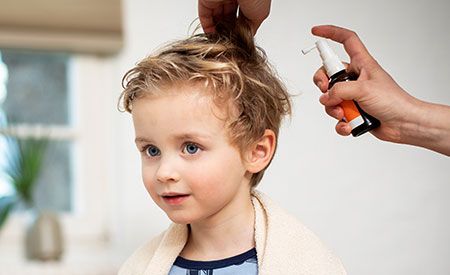 Ein kleines Kind mit blonden, leicht lockigen Haaren sitzt ruhig, während eine Person ein Läusespray auf sein Haar aufträgt. Das Kind trägt ein Handtuch über den Schultern.