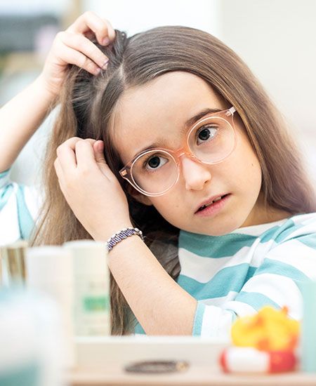 Ein Mädchen mit Brille untersucht sorgsam ihre Kopfhaut, indem es eine Haarsträhne anhebt. Sie trägt ein gestreiftes Shirt und sitzt an einem Tisch mit verschiedenen Gegenständen.