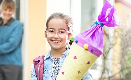Ein fröhliches Mädchen mit Brille und Schulranzen hält eine bunt dekorierte Schultüte. Im Hintergrund steht eine Frau lächelnd in der Tür eines Hauses.