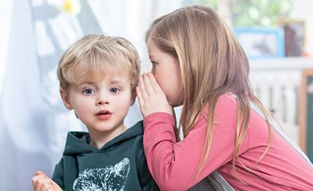 Ein blondes kleines Kind in einem dunkelgrünen Pullover schaut erstaunt, während ein Mädchen mit langen braunen Haaren ihm ins Ohr flüstert. Beide Kinder sitzen in einem hellen Raum.