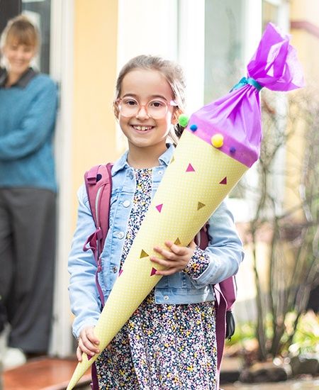 Ein fröhliches Mädchen mit Brille und Schulranzen hält eine bunt dekorierte Schultüte. Im Hintergrund steht eine Frau lächelnd in der Tür eines Hauses.