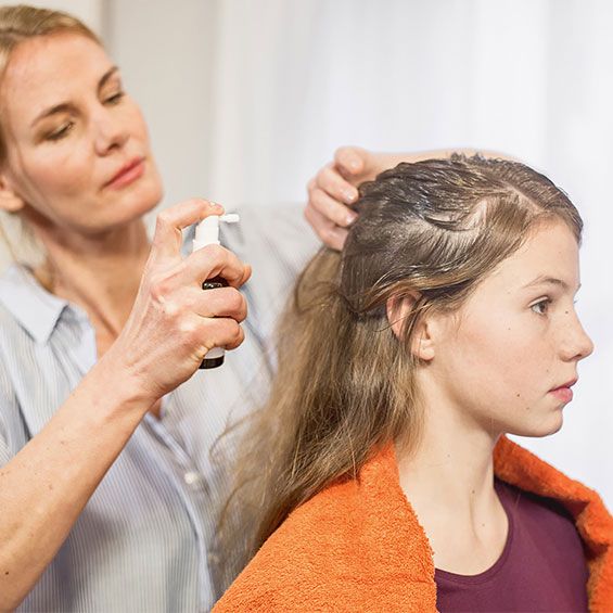 Eine Frau sprüht NYDA® Läusespray in das Haar eines Mädchens, das ein orangefarbenes Handtuch über den Schultern trägt. Das Mädchen sitzt ruhig und lässt die Behandlung durchführen.