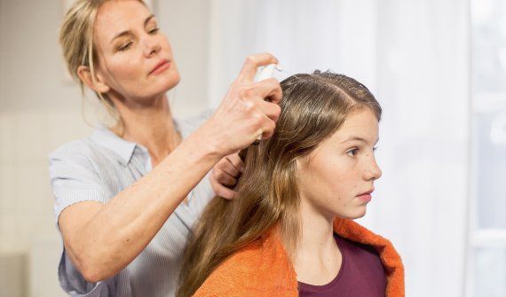 Eine Frau sprüht einem Mädchen NYDA® Läusespray auf die Kopfhaut, während diese ruhig dasitzt und ein Handtuch über den Schultern trägt.