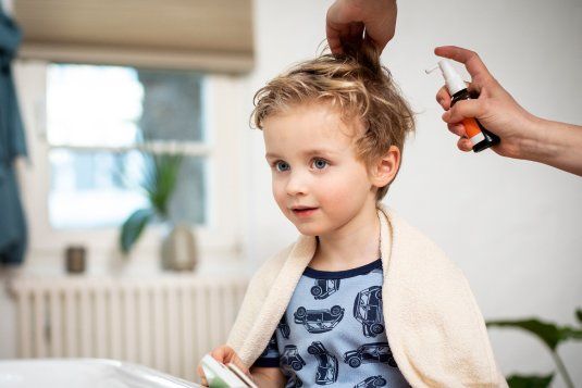 Ein kleines Kind mit blonden, leicht lockigen Haaren sitzt ruhig, während eine Person ein Läusespray auf sein Haar aufträgt. Das Kind trägt ein Handtuch über den Schultern.